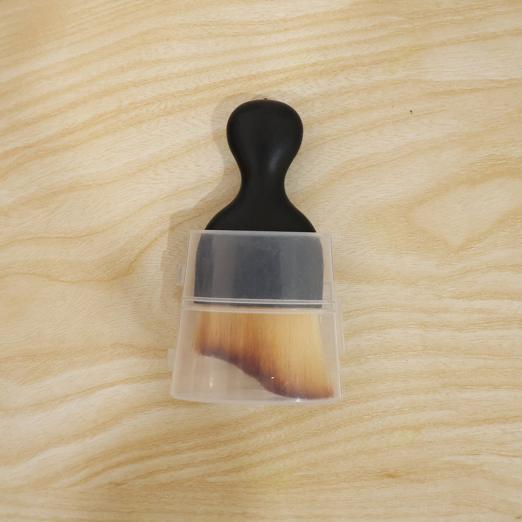 Mini cleaning brush with black handle, tan bristles, and clear protective cover on wood