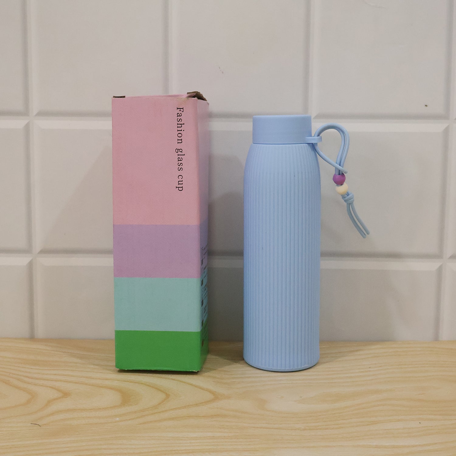 Light blue ribbed glass water bottle with silicone lid and carry loop beside product box