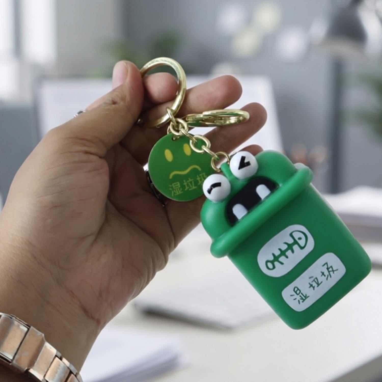 Hand holding green mini trash can keychain charm with gold split rings and chain