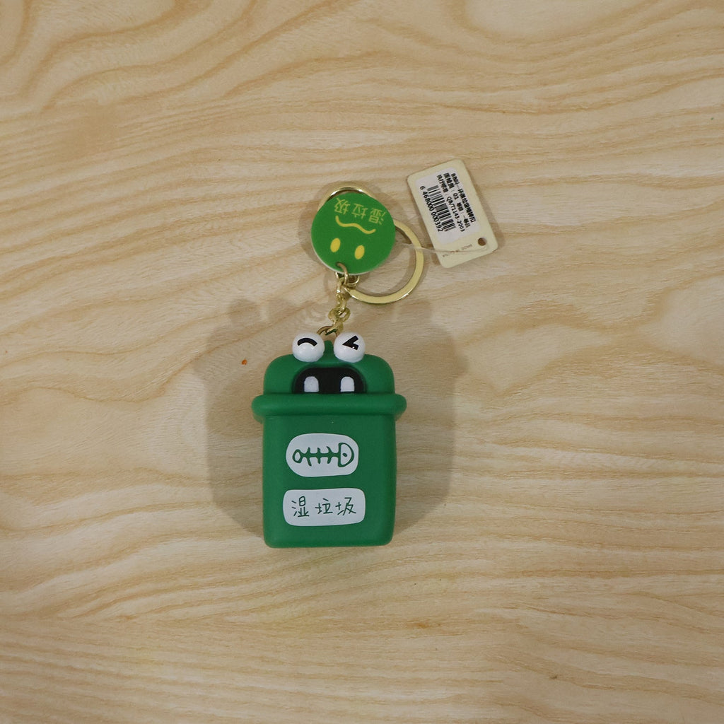 Green mini trash can charm keychain with gold rings and white tag on wooden surface