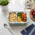 Two-compartment stainless lunch box with transparent lid, mint base, and spoon