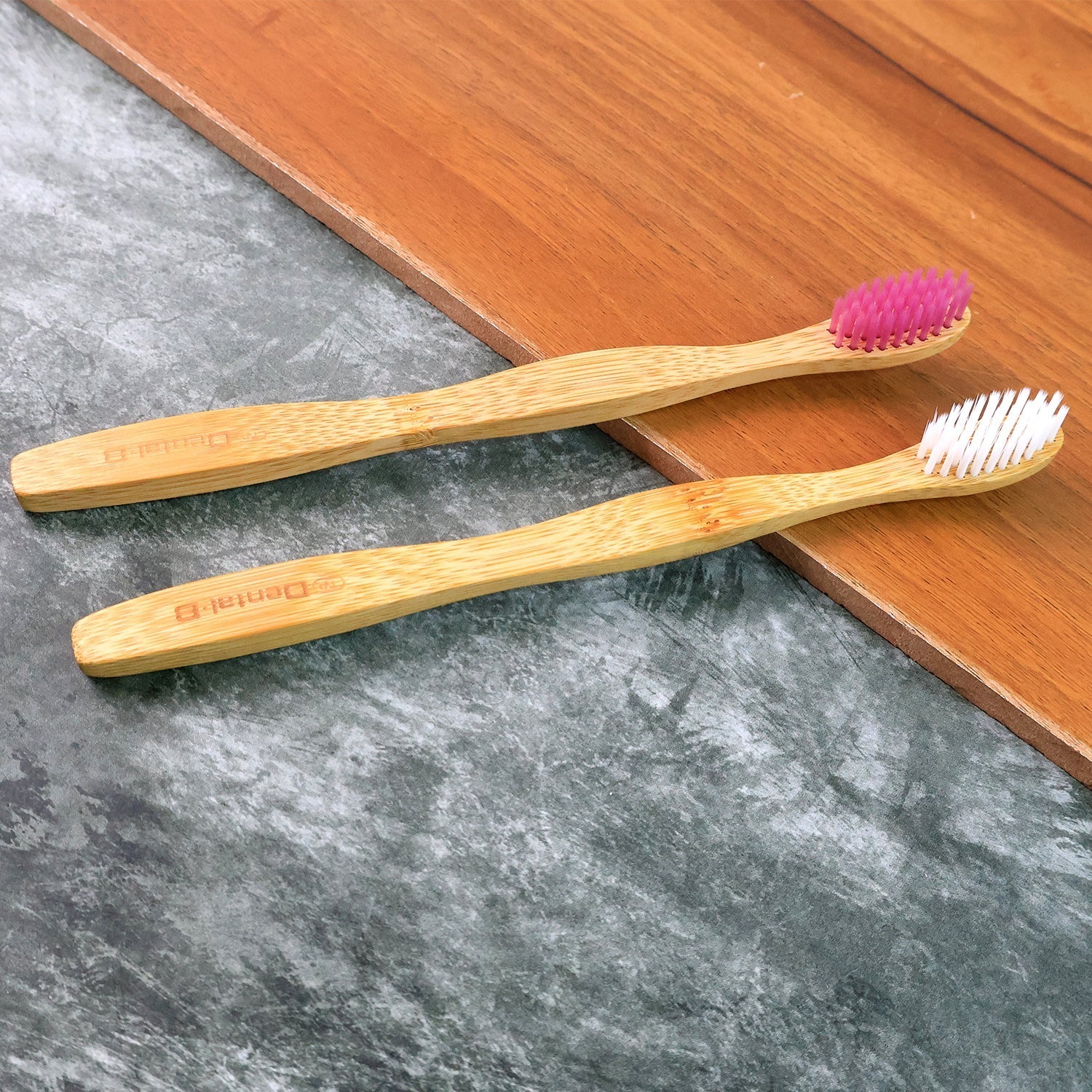 Two bamboo toothbrushes with soft bristles—white and purple—on a wooden board